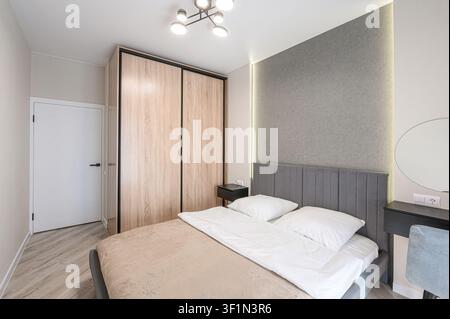 chambre moderne dotée d'un lit avec des draps blancs, une tête de lit grise et une couette beige. Une armoire en bois, une table de chevet et une coiffeuse avec un M rond Banque D'Images