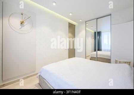 Intérieur de chambre propre et lumineux. Des murs blancs, une armoire à miroir et un lit avec une couette blanche dominent la scène. Une horloge dorée est accrochée au mur Banque D'Images