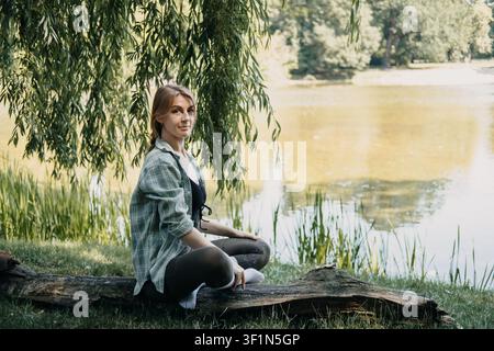 Jeune femme assise sur un arbre tombé au bord du lac, pause contemplative, échappatoire sereine, style de vie axé sur les soins personnels, retraite en solo Banque D'Images