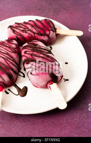Trois popsicles à base de fraises, de bananes, de betteraves et de yaourt grec sont posées sur une assiette. Ils sont arrosés de chocolat et dégustés pendant un d ensoleillé Banque D'Images
