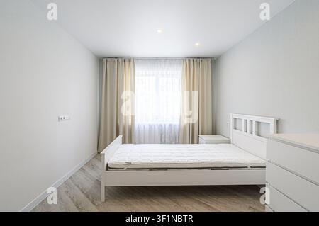 chambre minimaliste lumineuse avec des murs blancs et un lit blanc. Des rideaux encadrent une fenêtre, et une commode se trouve dans le coin. Lignes épurées et une palette neutre Banque D'Images