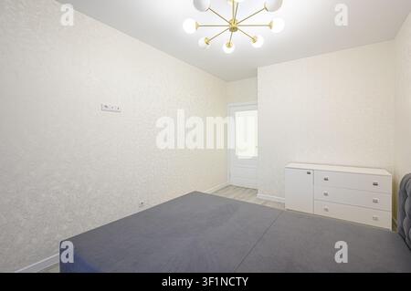 Chambre lumineuse dotée d'un lit gris, d'une commode blanche, de murs à motifs, d'un plafonnier moderne et d'une porte en arrière-plan Banque D'Images