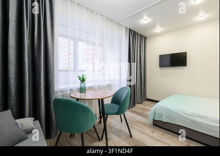 un appartement studio lumineux comprend un petit coin repas près d'une fenêtre avec des chaises vertes et un lit avec une télévision murale Banque D'Images