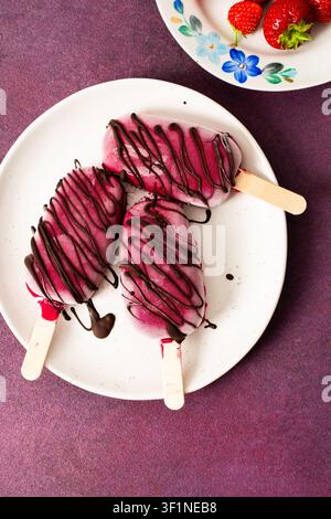 Trois popsicles à base de fraises, de bananes, de betteraves et de yaourt grec sont posées sur une assiette. Ils sont arrosés de chocolat et dégustés pendant un d ensoleillé Banque D'Images