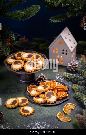 Biscuits de Noël avec un motif yin yang sur un support à gâteau et une assiette en métal sombre, entourés d'oranges séchées, de pommes de pin et de branches de sapin festives Banque D'Images