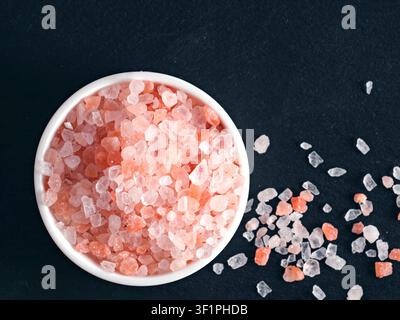Sel rose himalayen grossier dans un petit bol en céramique blanche sur un fond d'ardoise noire. Composition en vue plongeante à angle élevé avec des détails nets et un espace de copie. Parfait pour la cuisine ou le bien-être Banque D'Images