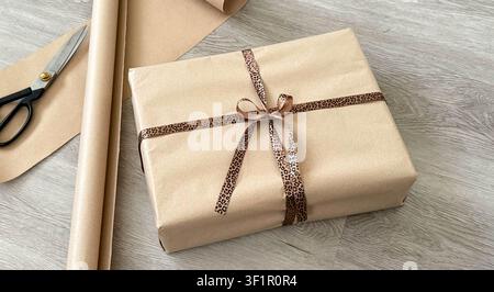 Coffret cadeau enveloppé d’un ruban à motif léopard posé sur une surface en bois, entouré de ciseaux et de rouleaux de papier cadeau dans un environnement intérieur bien éclairé Banque D'Images