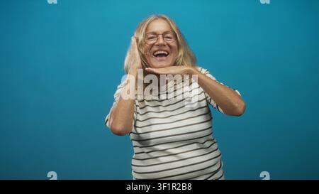 Femme senior riant avec les mains encadrant le visage, portant une chemise rayée et des lunettes, posant de manière ludique sur fond de studio bleu ; joie énergie ludique. Banque D'Images