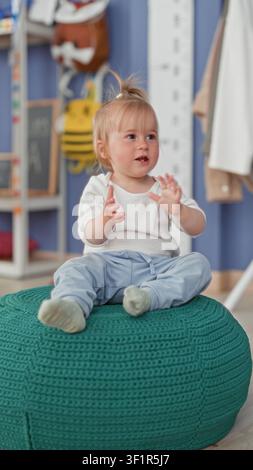 Petite fille blonde assise sur un pouf tricoté vert, frappant dans ses mains et souriant dans une salle de jeux pour enfants à l'intérieur d'un bâtiment ; émerveillement ludique. Banque D'Images