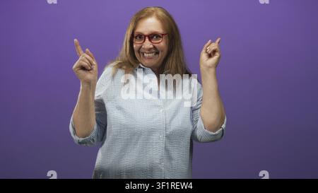 Sénior femme pointe ses deux index vers le haut et sourit largement, portant des lunettes rouges et une chemise à carreaux clairs en studio ; célébration de joie. Banque D'Images
