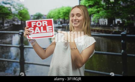 La femme pointe le doigt à pour le signe de vente sur la rampe de canal de rue tout en tenant l'écriteau blanc et souriant ; promotion de la confiance des ventes. Banque D'Images
