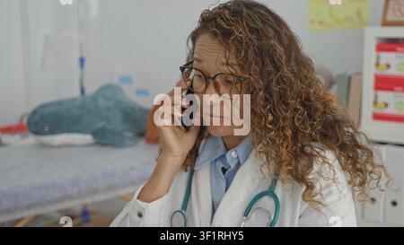 Femme d'âge moyen médecin en manteau blanc tenant smartphone à l'oreille, stéthoscope visible dans la salle d'examen clinique avec lit d'examen, jouet en peluche et boîtes médicales; Banque D'Images