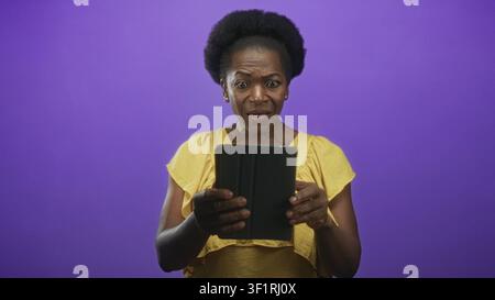 Femme tenant une tablette à deux mains, vêtue d'une robe jaune, regardant l'écran en studio ; curiosité inquiète. Banque D'Images