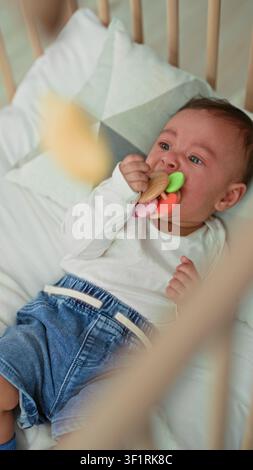 Garçon hispanique en bas âge dans le berceau mordant un anneau coloré de dentition avec la bouche et les yeux largués dans une pépinière de bâtiment ; inconfort de dentition. Banque D'Images