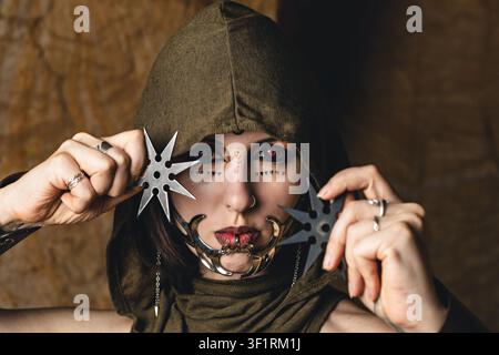 Portrait dramatique d'une femme dans un costume à capuche avec des lentilles de contact rouges, des bijoux de visage et des étoiles de lancer de style ninja, personnage de fantaisie sombre Hallowee Banque D'Images
