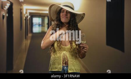 Femme adolescente hispanique tient une barre de crème glacée saupoudrée et presse la main contre la poitrine tout en grignant dans un couloir de bâtiment ; préoccupation de nausées. Banque D'Images