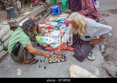 Deux hommes âgés locaux aux cheveux longs vendant des livres accroupis au bord de la route jouant aux échecs dans la rue à Yangon (Rangoun), Myanmar (Birmanie) Banque D'Images