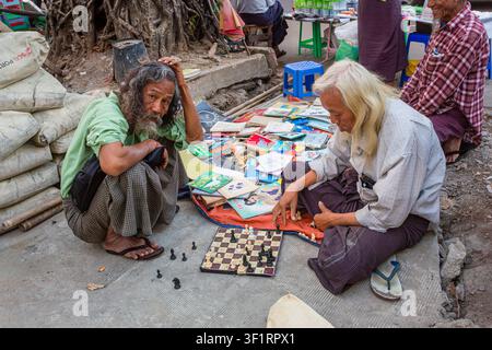 Deux hommes âgés locaux aux cheveux longs vendant des livres accroupis au bord de la route jouant aux échecs dans la rue à Yangon (Rangoun), Myanmar (Birmanie) Banque D'Images