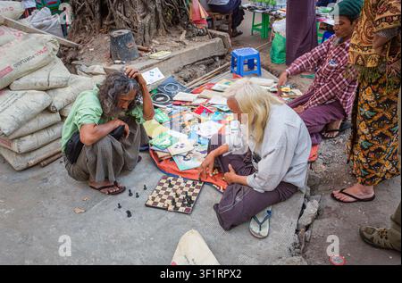 Deux hommes âgés locaux aux cheveux longs vendant des livres accroupis au bord de la route jouant aux échecs dans la rue à Yangon (Rangoun), Myanmar (Birmanie) Banque D'Images
