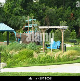 Panama City, FL, USA. 14 juil. 2026. Une balancelle de porche ombragée par une pergola et des plantations d’herbe indigène se trouvent devant l’aire de jeux en forme de navire pirate et l’abri de pique-nique du Millville Waterfront Park sur Watson Bayou lors de la cérémonie de coupure de ruban marquant la réouverture officielle du parc. La rénovation financée par la Community Redevelopment Agency – amphithéâtre, sentier de marche éclairé, aire de jeux, abris de pique-nique et balancelles au bord de l’eau – a été achevée avec environ 200 000 $ de moins que le budget, faisant partie de l’effort de réinvestissement dans les parcs de Panama City dans le quartier historique des moulins durement touché par l’ouragan Michael en 2018. Crédit : Tosh Persons / Alamy Live News Banque D'Images