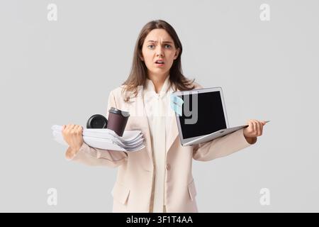 Jeune femme d'affaires submergée tenant une pile de papiers, un café et un ordinateur portable avec une note de délai isolée sur un fond clair Banque D'Images
