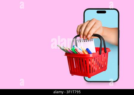 Main tenant un panier d'achat rouge avec des fournitures scolaires émergeant d'un écran de smartphone, éducation en ligne et retour à l'école Banque D'Images