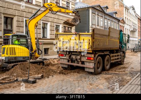 Excavateur jaune chargeant du sable dans le plateau d'un camion-benne pendant des travaux de construction routière sur une rue de la ville Banque D'Images