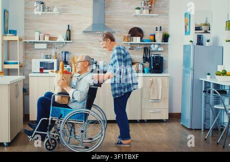 Une vieille femme pousse son mari senior invalide dans un fauteuil roulant après être revenue des courses au supermarché avec un sac en papier, dans une routine pratique de cuisine. Banque D'Images