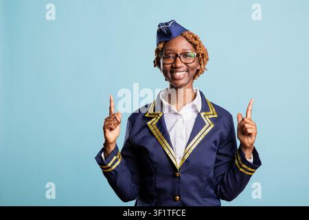 Hôtesse de l'air heureuse en uniforme bleu pointe vers l'espace vide contre un fond bleu. L'hôtesse de vol sourit et gesticule avec les index, promouvant le marketing aérien et les concepts de voyage. Banque D'Images