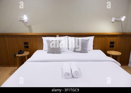 Un lit d'hôtel moderne avec des draps blancs et des oreillers gris soigneusement disposés sur une tête de lit en bois, des serviettes roulées placées sur la couette transmettant le calme et l'accueil Banque D'Images