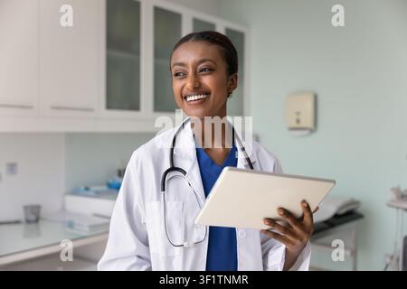 Jeune médecin noire joyeuse tenant une tablette dans un bureau de clinique Banque D'Images