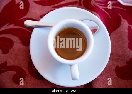 Tasse à expresso reposant sur une soucoupe sur un tissu à motif rouge dans un cadre de culture du café détendu, vue de dessus avec des matériaux en céramique et métalliques, blanc et Banque D'Images