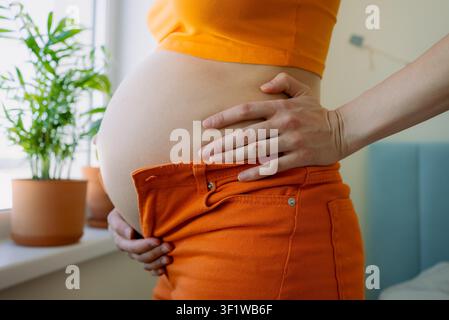 Femme enceinte debout, montrant son ventre qui grossit, luttant pour fermer son jean orange serré autour d'un abdomen en expansion, illustrant la transformation physique pendant la grossesse Banque D'Images