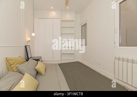Chambre lumineuse avec armoires encastrées blanches et étagères ouvertes, un lit avec une parure grise et des coussins moutarde, des suspensions et un radiateur blanc. Banque D'Images