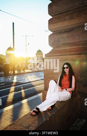 Femme élégante se détendant sur un pont historique de la ville au coucher du soleil, profitant de l'heure dorée dans une tenue estivale à la mode et des lunettes de soleil. Banque D'Images