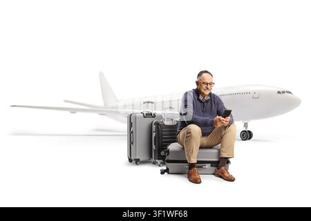Homme mature assis sur une valise devant un avion et utilisant un smartphone isolé sur fond blanc Banque D'Images
