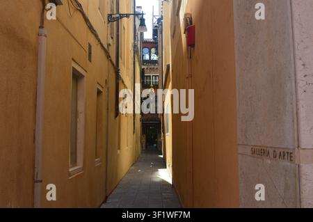 Ruelle vénitienne étroite et ensoleillée bordée de murs jaune chaud menant vers une enseigne de galerie d'art à Venise, Italie Banque D'Images
