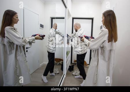 Une personne se tient devant un miroir en utilisant un sèche-cheveux. Une autre personne ajuste sa tenue à proximité. Un chat est assis au sol en arrière-plan. Le set Banque D'Images