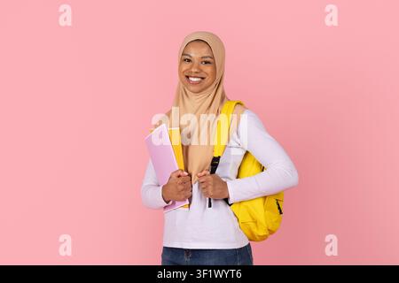 Étudiante musulmane noire portant un hijab et tenant des cahiers colorés ainsi qu'un sac à dos jaune Banque D'Images