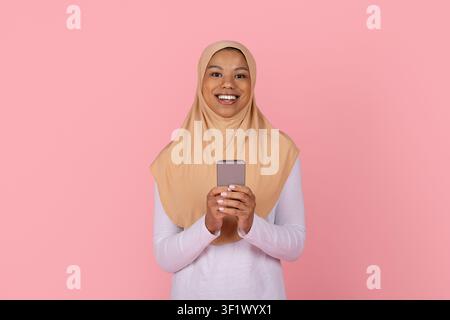 Jeune femme musulmane noire joyeuse portant un hijab beige et tenant un smartphone dans les mains Banque D'Images