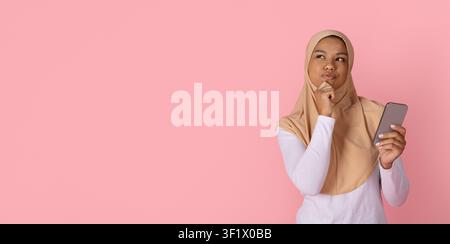 Femme musulmane noire pensive portant un hijab beige, tenant un smartphone et touchant son menton Banque D'Images