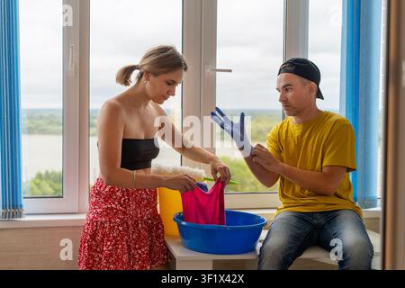 Jeun couple nettoyant l'appartement ensemble, femme lavant les fenêtres tandis que l'homme enfile des gants de protection en caoutchouc. Ménage, travail d'équipe, hygiène domestique, Banque D'Images