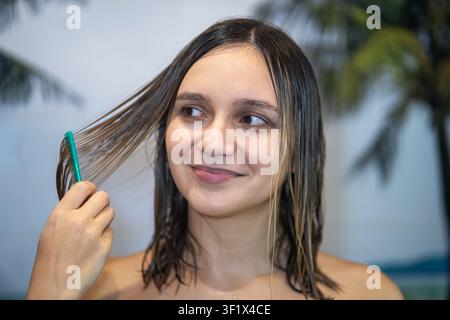 Jeune femme peignant ses cheveux mouillés après la douche, souriant tout en prenant soin de ses cheveux sains. Routine de beauté quotidienne, soins des cheveux, soins personnels, bien-être et wellness a Banque D'Images
