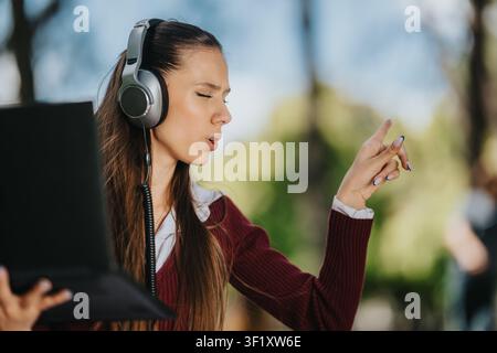 Jeune femme portant des écouteurs chantant tout en utilisant un ordinateur portable en plein air dans un parc Banque D'Images