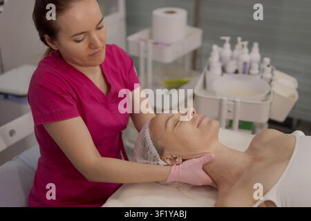 Cosmétologue effectue un massage du cou et du visage sur une femme âgée dans un institut de beauté, offrant des services professionnels de soins de la peau axés sur la relaxation Banque D'Images