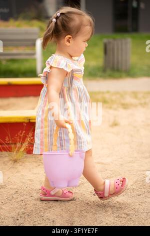Profil latéral d'un tout-petit en robe rayée pastel avec des couettes marchant sur un terrain de jeu sablonneux à midi, portant un seau lavande, scène d'été spontanée et chaleureuse Banque D'Images