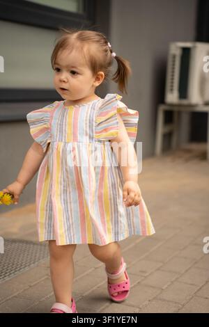 Petite fille en robe à rayures pastel et sandales roses marchant sur un trottoir pavé tenant de petites fleurs jaunes, portrait spontané en plein air avec une lumière naturelle douce Banque D'Images