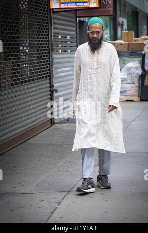 Un homme musulman grand et mince portant une chemise blanche longue et un chapeau kufi vert marchant dans le quartier Little Bangladesh de Jackson Heights, Queens, New York. Banque D'Images