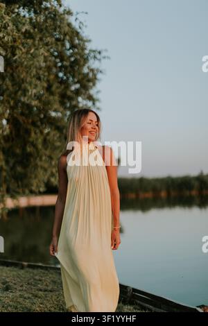 Une femme se tient près de l'eau avec de la haute herbe autour d'elle. Elle sourit doucement dans une robe légère alors que le soleil se couche en arrière-plan. Le ciel a une lueur chaude Banque D'Images