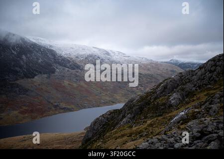 Des sommets enneigés encadrent un lac calme et profond. Des ciels gris pèsent bas sur des pentes escarpées et moussues. Le givre murmure le long des arêtes déchiquetées, évoquant le froid. Calme Banque D'Images